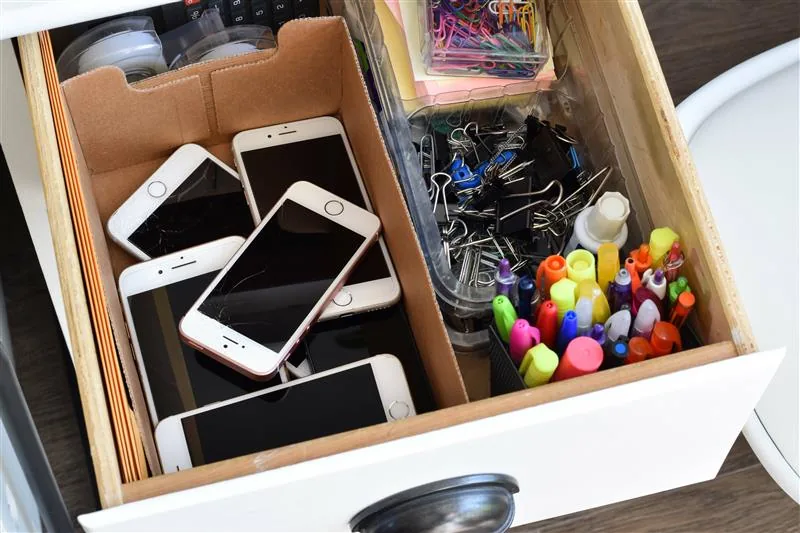 Staršie modely iPhonov uložené v zásuvke pripravené na výkup iPhonu. V zásuvke sú aj kancelárske potreby - farebné perá a papieriky, klipy a iné drobnosti.